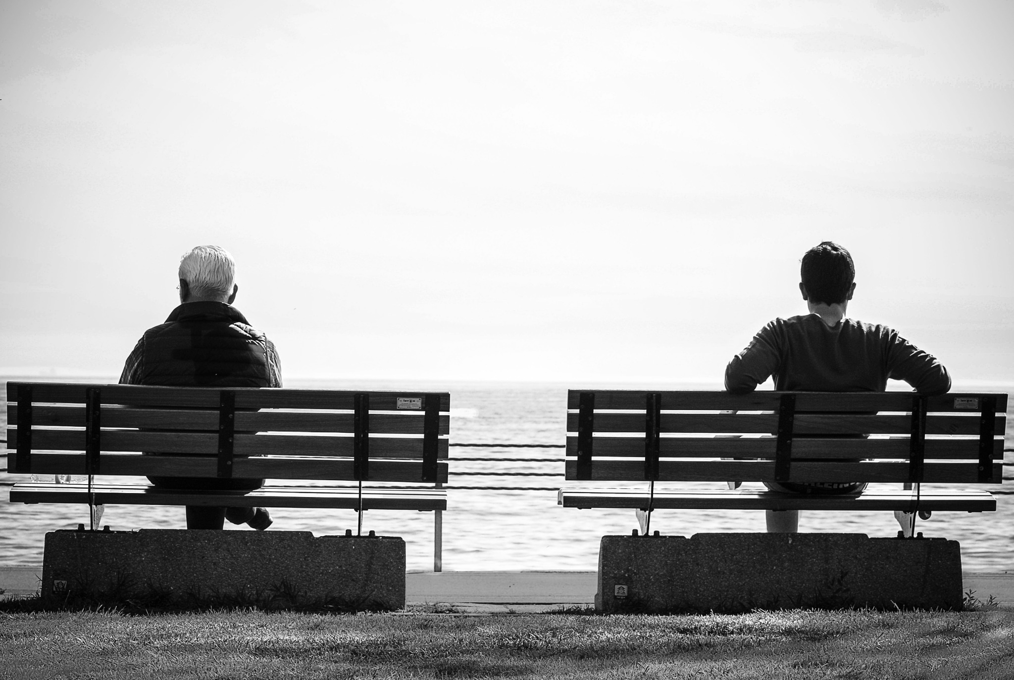 Two people sitting apart on separate benches facing the water